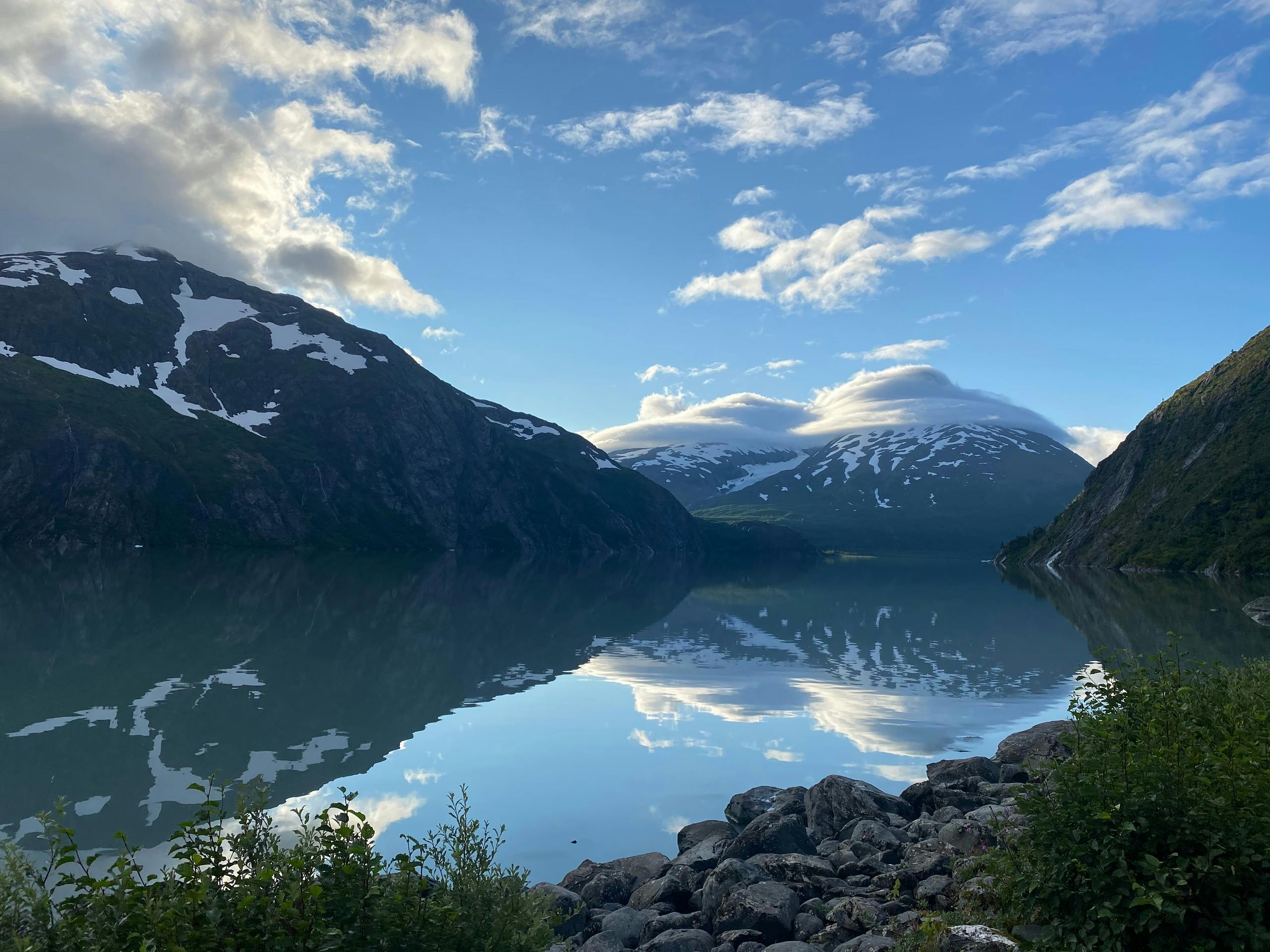 A serene mountain lake surrounded by snow-capped peaks under a partly cloudy blue sky, with reflections on the water.