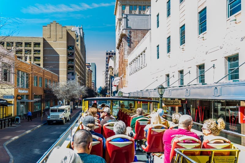 Pessoas a passear no andar superior de um autocarro de turismo por uma rua da cidade ladeada de edifícios sob um céu azul límpido.