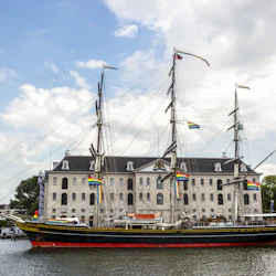 Ett stort segelfartyg med flera master och flaggor dockade framför en historisk stenbyggnad, under en delvis molnig himmel.