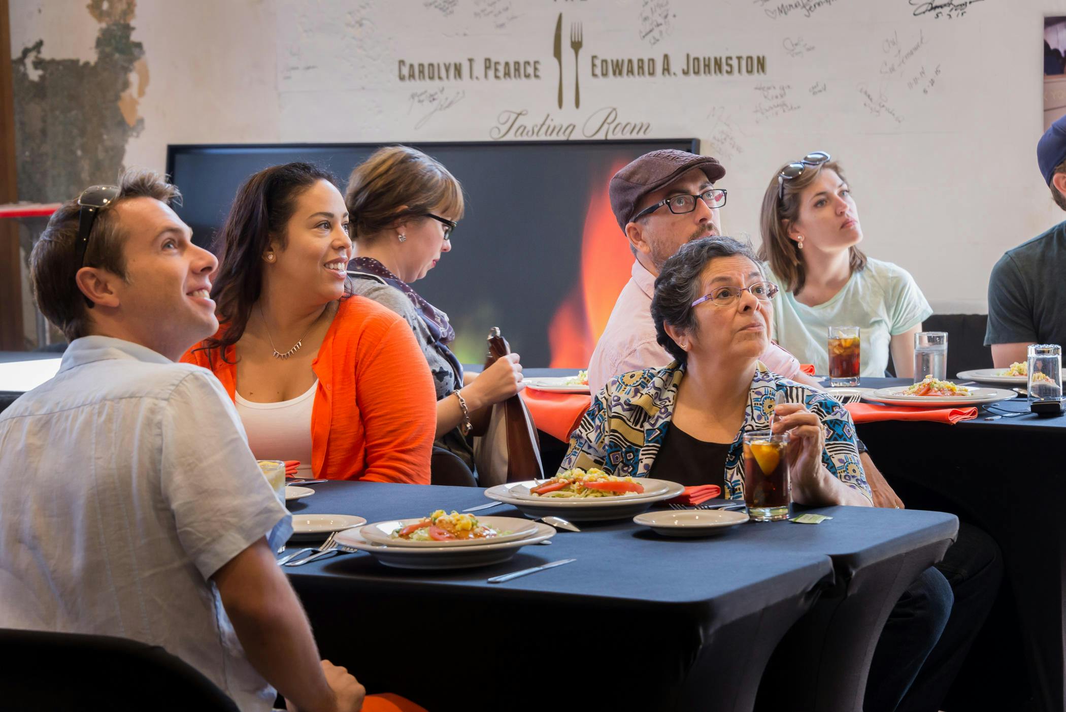 Een groep mensen rond tafels met eten en drinken, aandachtig luisterend in een ruimte met het label "Proeflokaal".