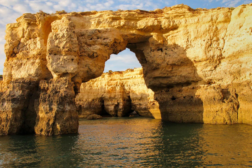 Natural rock arch over calm water, illuminated by golden sunlight, with clear sky in the background.