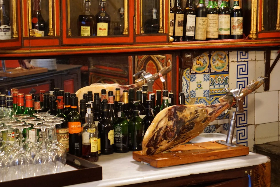 A bar counter displaying various liquor bottles, wine glasses, and a large leg of cured ham on a wooden stand.