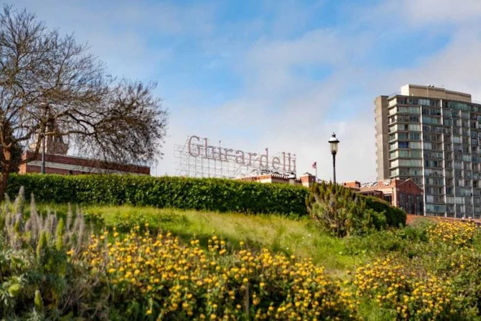 Ghirardelli sign seen over a grassy hill with yellow flowers, a lamppost, tree, and nearby buildings under a blue sky.