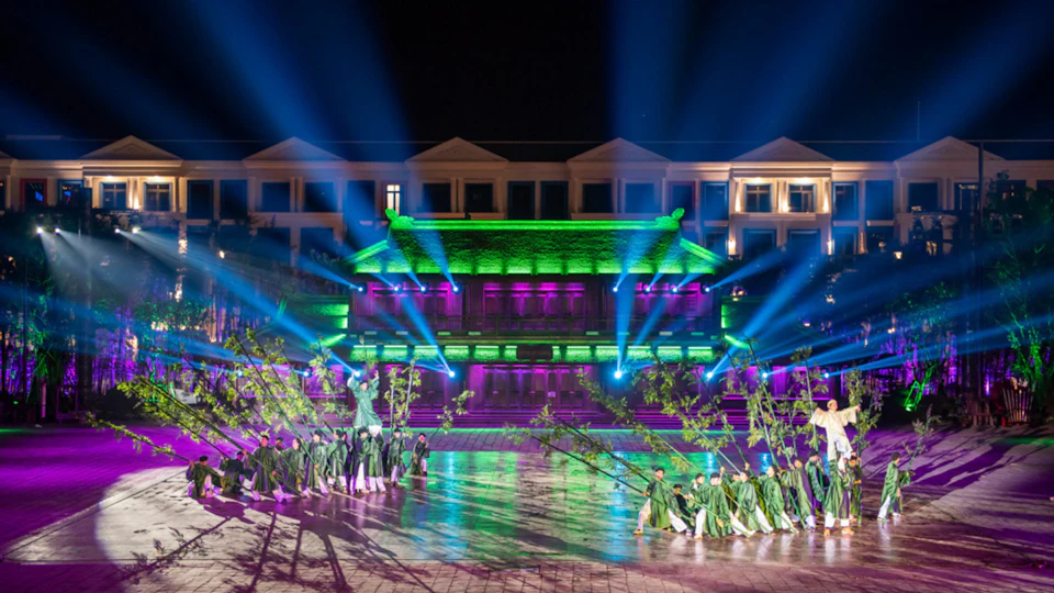 A vibrant stage performance featuring dancers in green costumes under colorful lights in front of a lit-up traditional building.