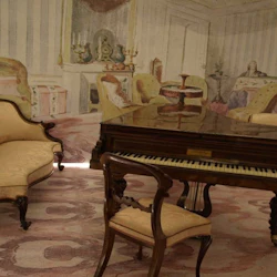 Elegant rum med ett flygel, en utsmyckad svimningssoffa och en väggmålning som visar en mysig interiörscen.