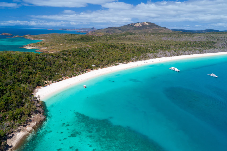 Aerial view of a sandy beach with clear turquoise water, surrounded by lush green hills and boats anchored near the shore.