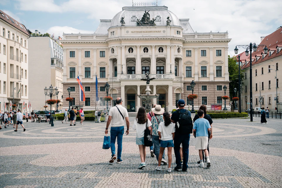 A group of people walk towards a large, ornate building with statues and flags in the foreground of a cobblestone plaza.