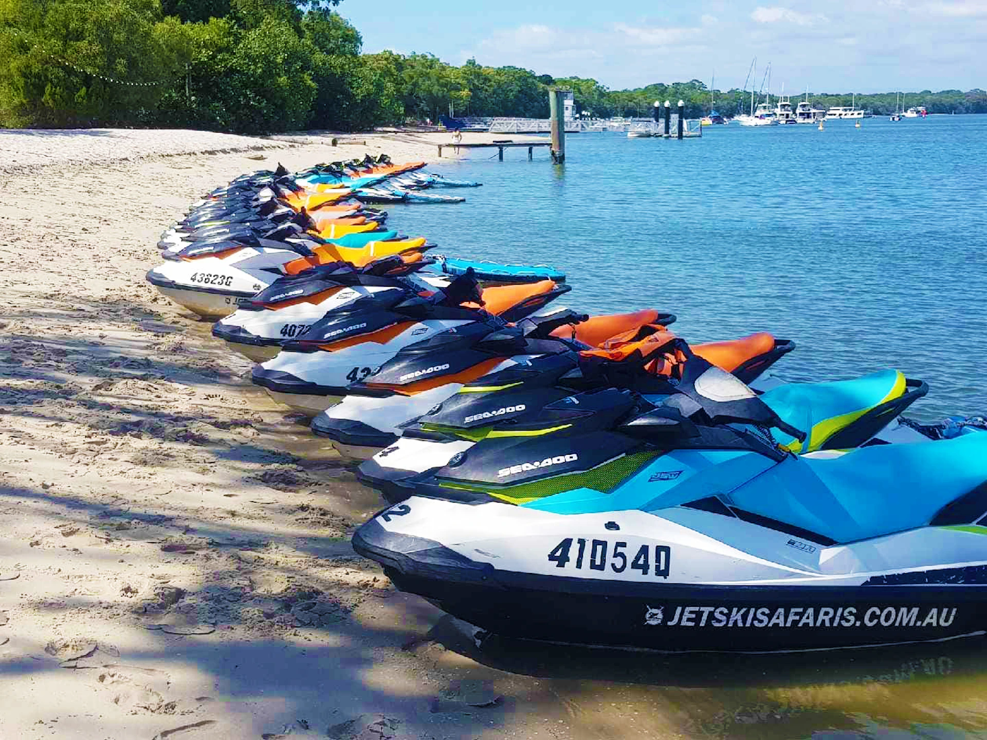A line of colorful jet skis is parked on a sandy beach next to a calm body of water, with trees and a dock in the background.