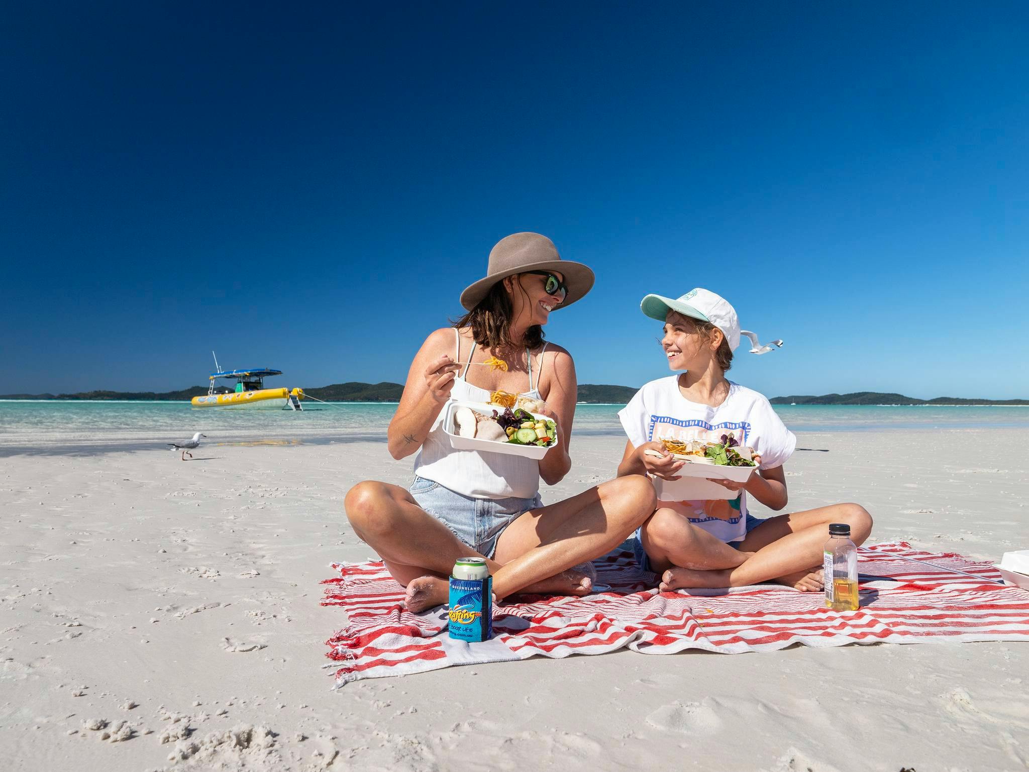 Two individuals sitting on a striped blanket on a sandy beach, eating from food containers under a clear blue sky.