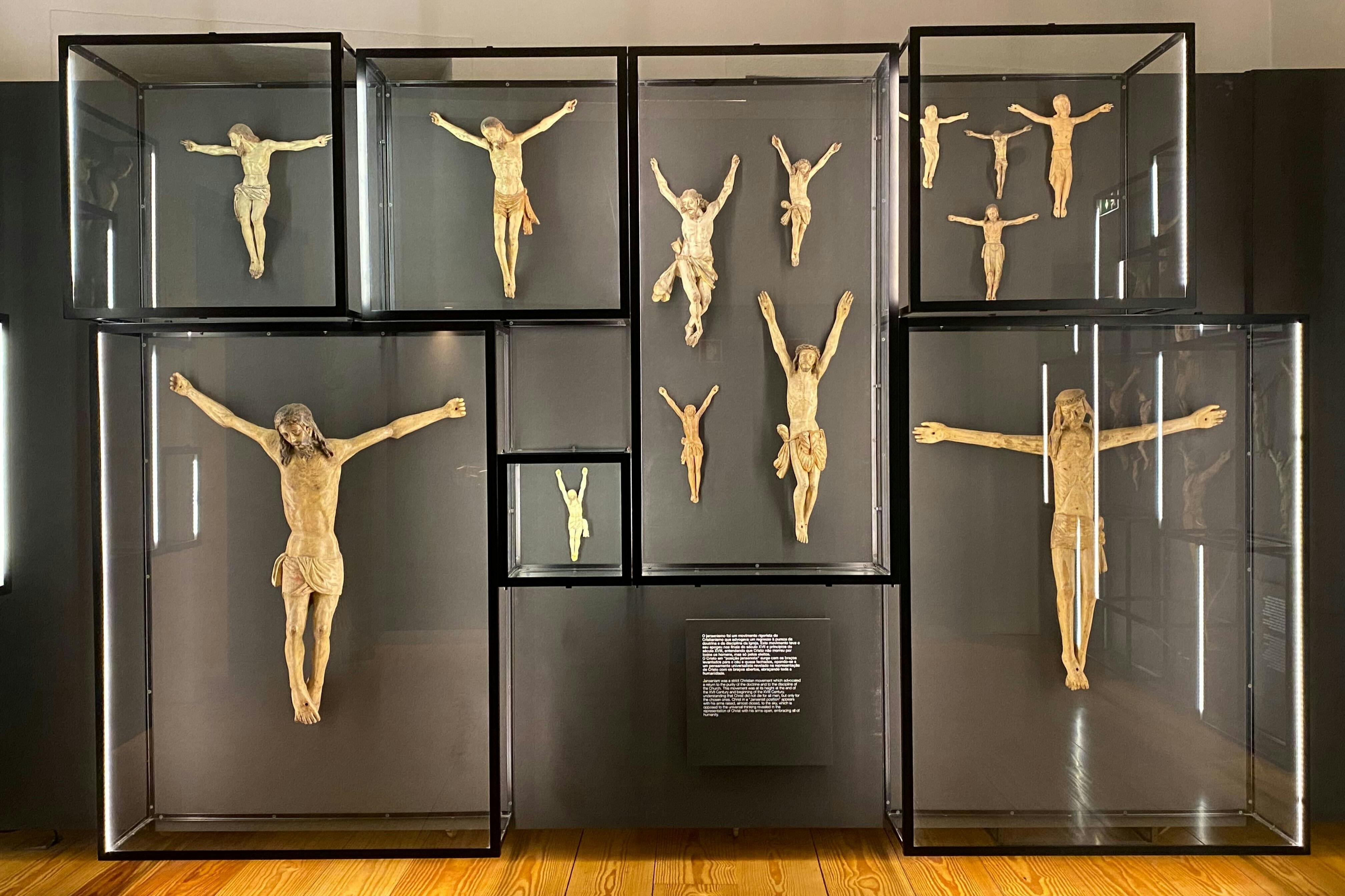 Různé krucifixy ve skleněných vitrínách uspořádaných na stěně, s popisnou plaketou uprostřed dole.
