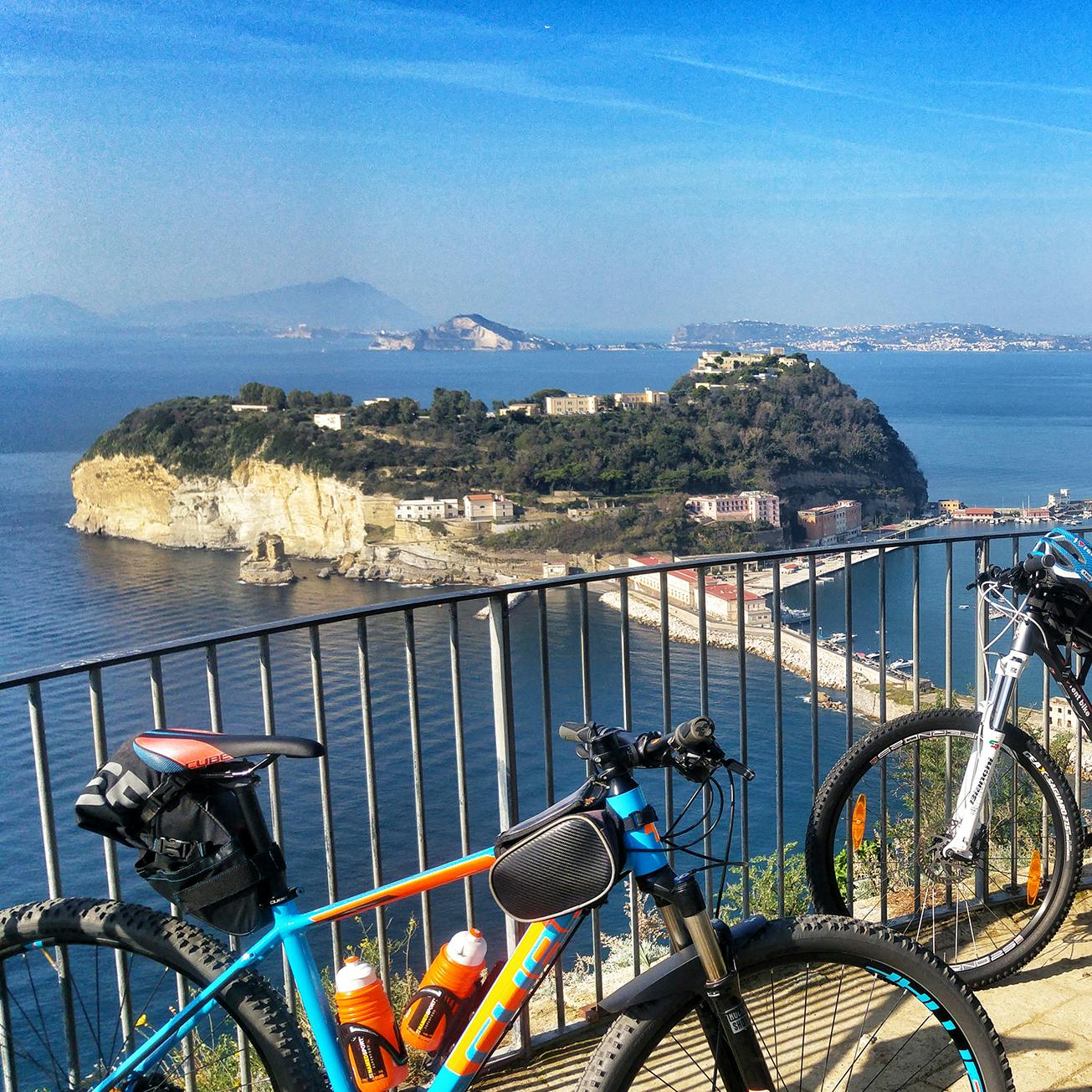 Due biciclette parcheggiate vicino a una ringhiera di metallo che si affaccia su un paesaggio costiero isolano con edifici, colline e cielo azzurro.