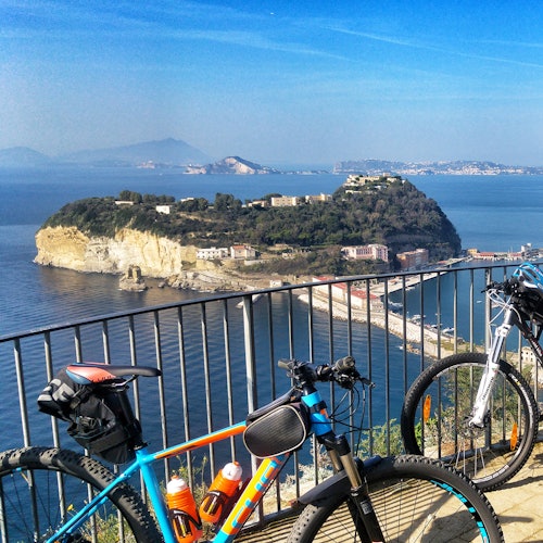 Two bicycles parked by a metal railing overlooking a coastal island landscape with buildings, hills, and blue sky.