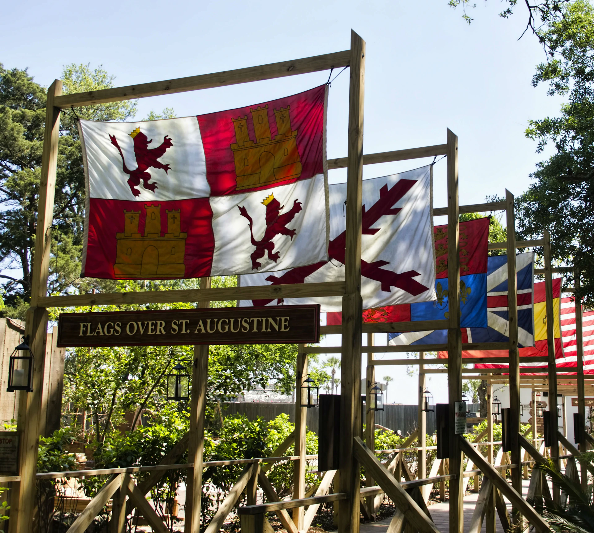 Colorful historic flags hang outdoors on wooden frames with a sign below reading "Flags Over St. Augustine." Trees and foliage surround the area.