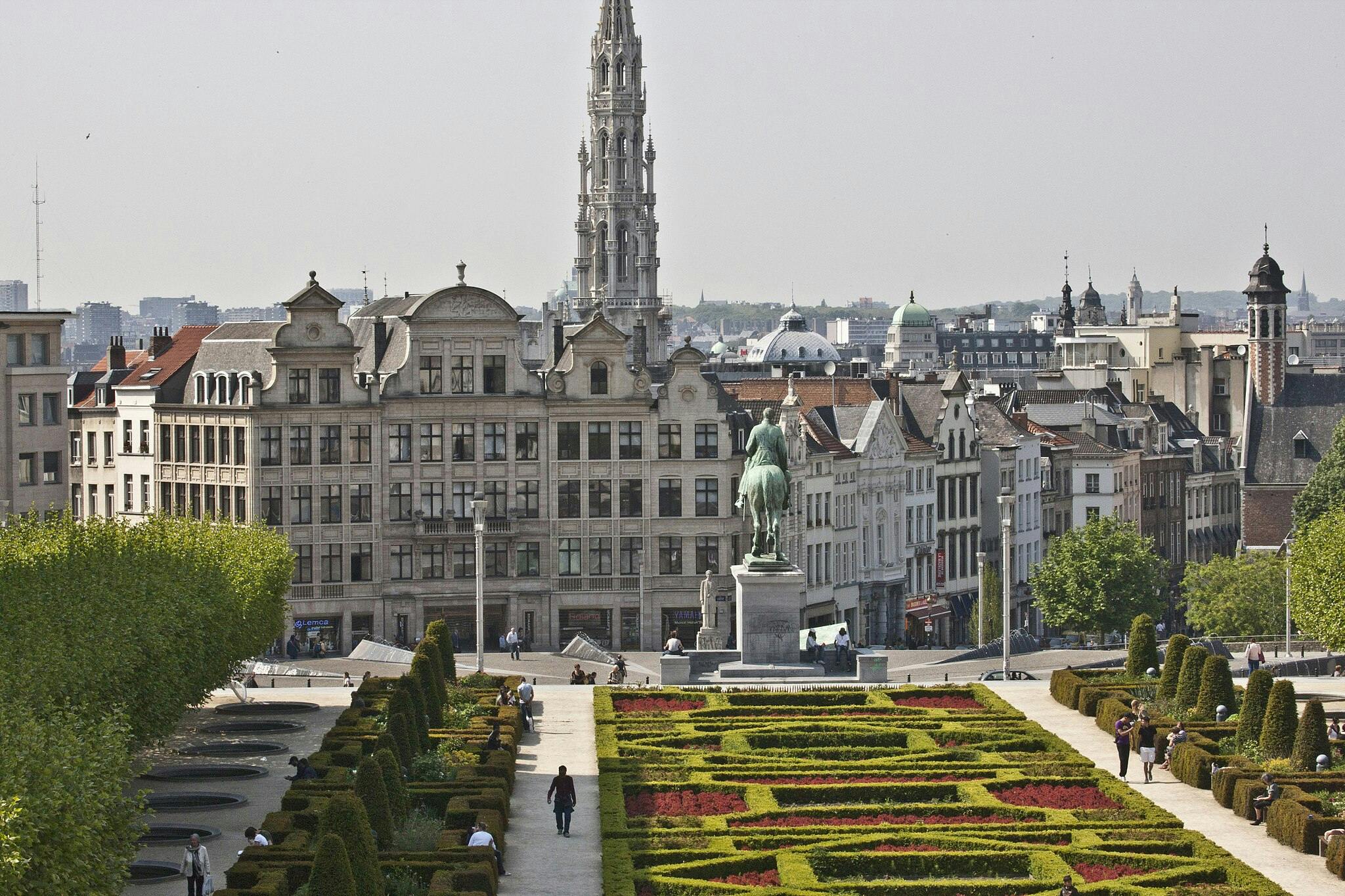 Well-manicured garden with hedges and flowers, people walking, statue of a rider on horseback, historic buildings in the background.