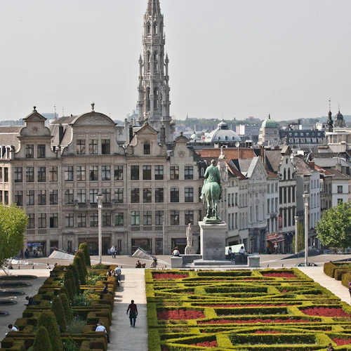 Välskött trädgård med häckar och blommor, människor som går, staty av en ryttare till häst, historiska byggnader i bakgrunden.