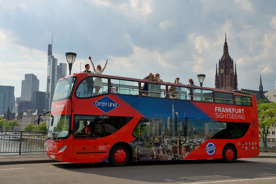 Bright red double-decker sightseeing bus with people on the top deck, surrounded by city buildings and lampposts.