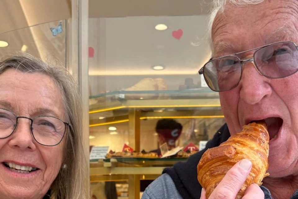 Two people sitting in front of a pastry shop, one eating a croissant and the other smiling.