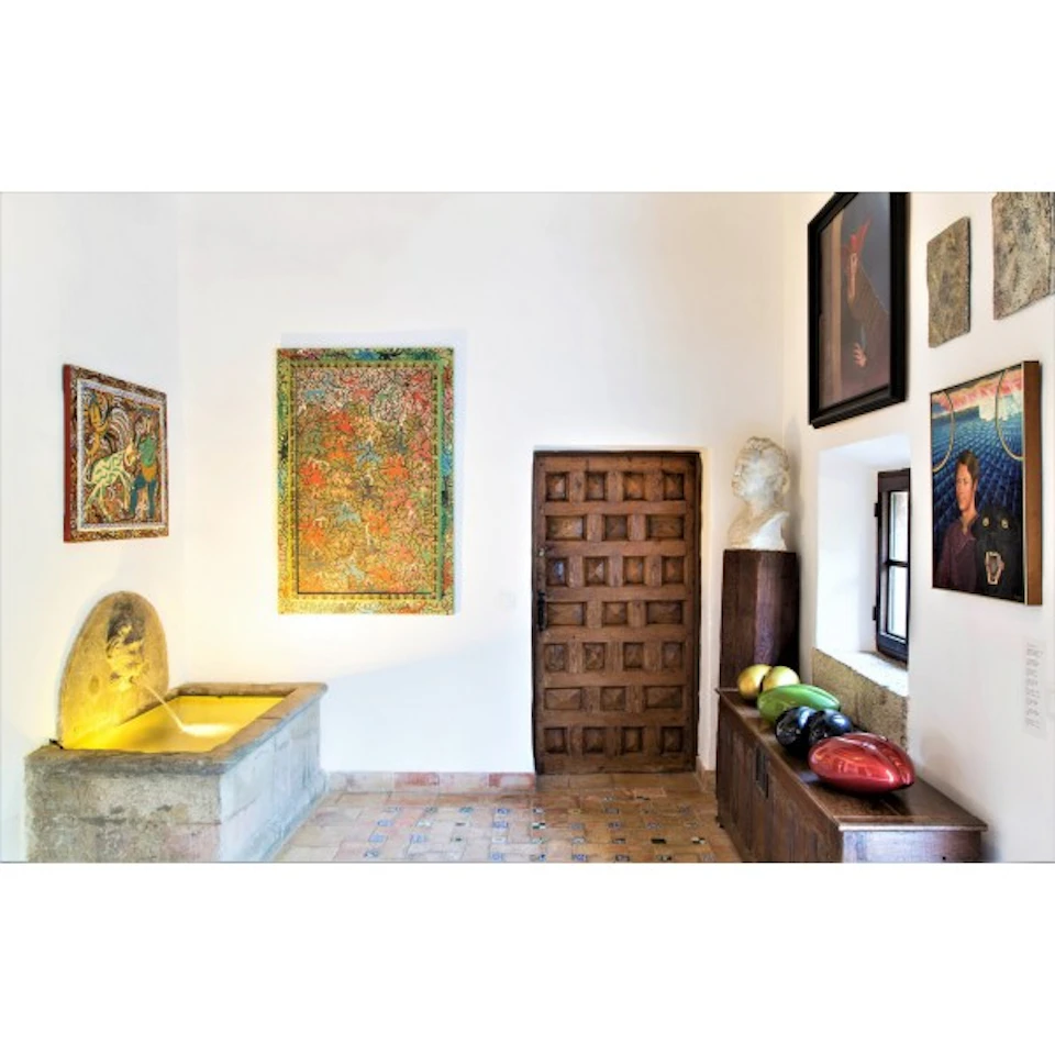 Room with tiled floor and wooden door, featuring colorful paintings, a stone sink, a bust on a stand, and ceramic fruit on a table.