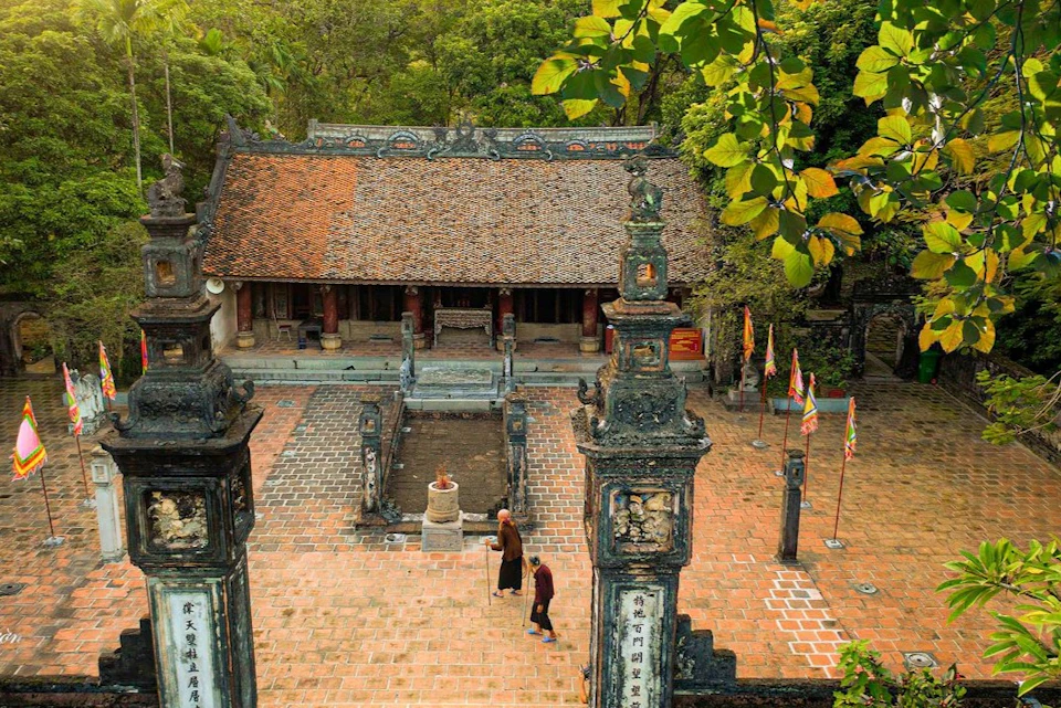 Aerial view of a historical courtyard with two people walking, ornate pillars, flags, and a tiled-roof building surrounded by trees.