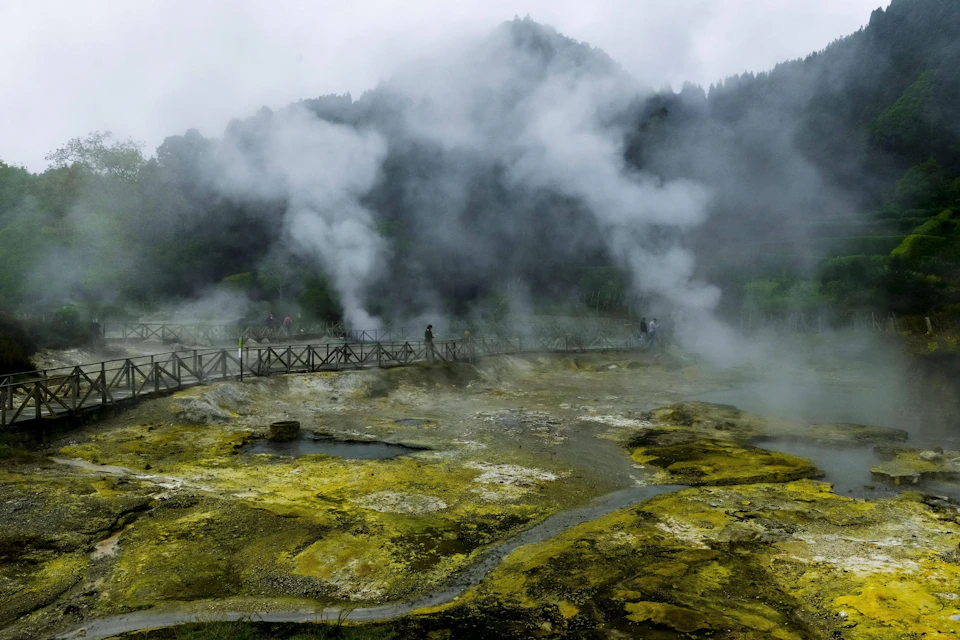 Foggy scene with steam rising from yellow-tinted ground. A few people walk across a wooden bridge, with misty trees in the background.