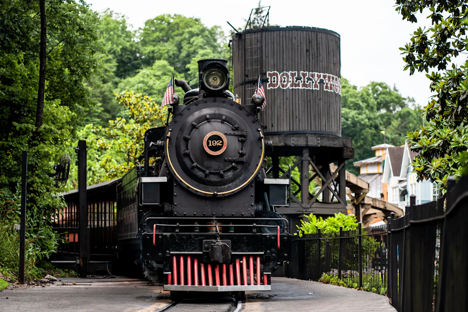 A vintage steam locomotive labeled "102" in front of a wooden water tower with "Dollywood" written on it, surrounded by greenery.