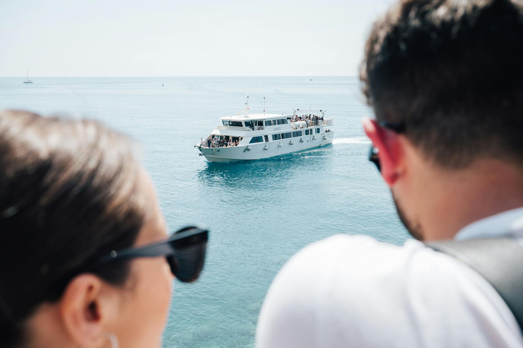 一名男子拿着双筒望远镜看着一艘白色渡船，船上满载着在平静海面上航行的乘客。