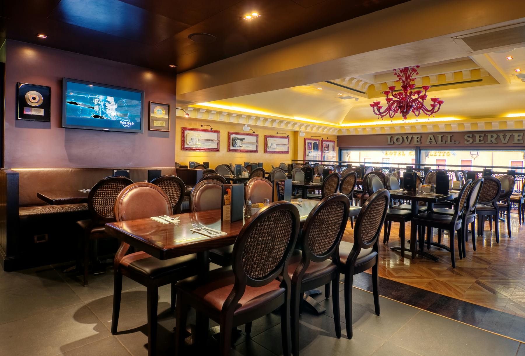 A well-lit restaurant with wooden tables, brown chairs, wall-mounted TV, framed pictures, and a pink chandelier.