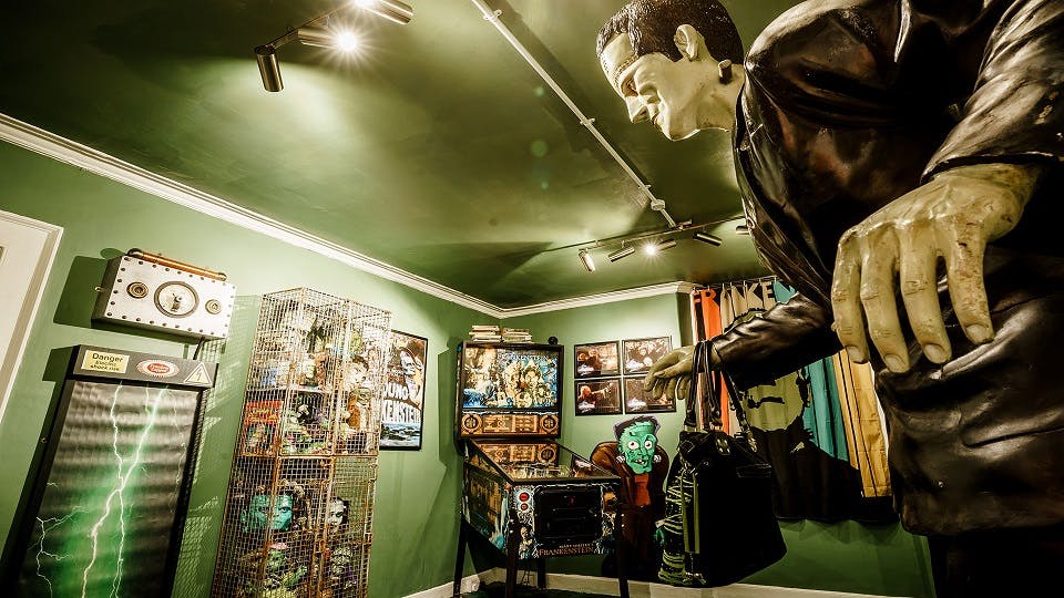 A green brightly lit room with Frankenstein related paraphilia including pinball machine and large monster statue 