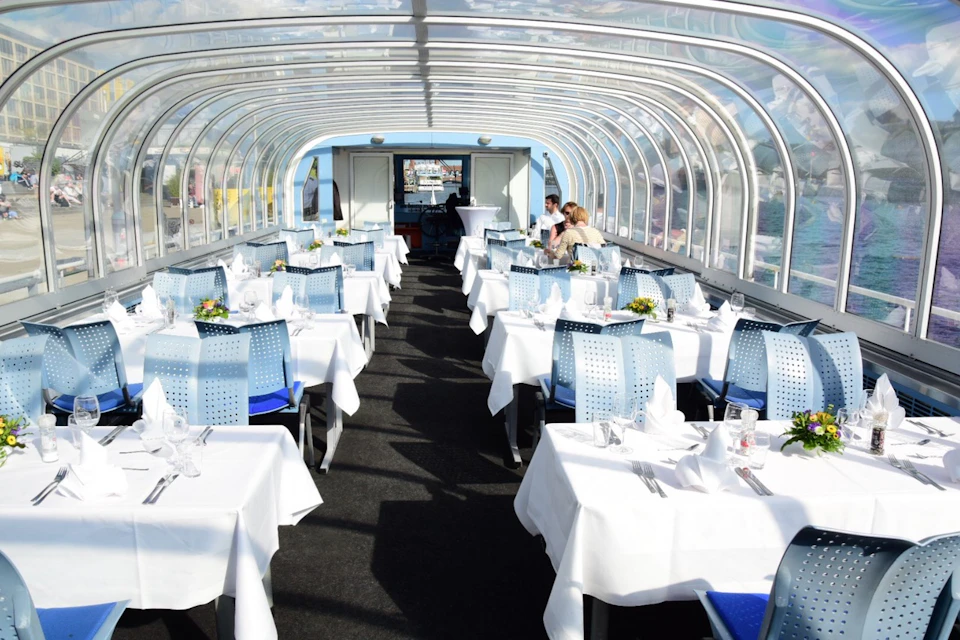 Restaurant with glass roof, neatly set tables with white linens, blue chairs, two people dining in the background.