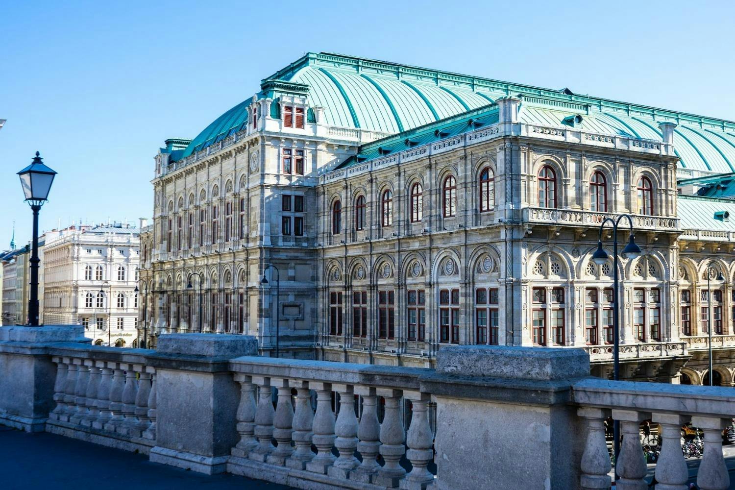 Historic building with ornate detailing and a green domed roof, viewed from behind a balustrade with a streetlamp on the left.