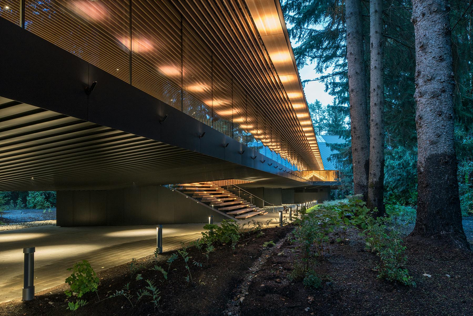 Bâtiment moderne avec une façade en verre et des lattes de bois, monté sur pilotis avec des escaliers éclairés, entouré d'arbres et de plantes.