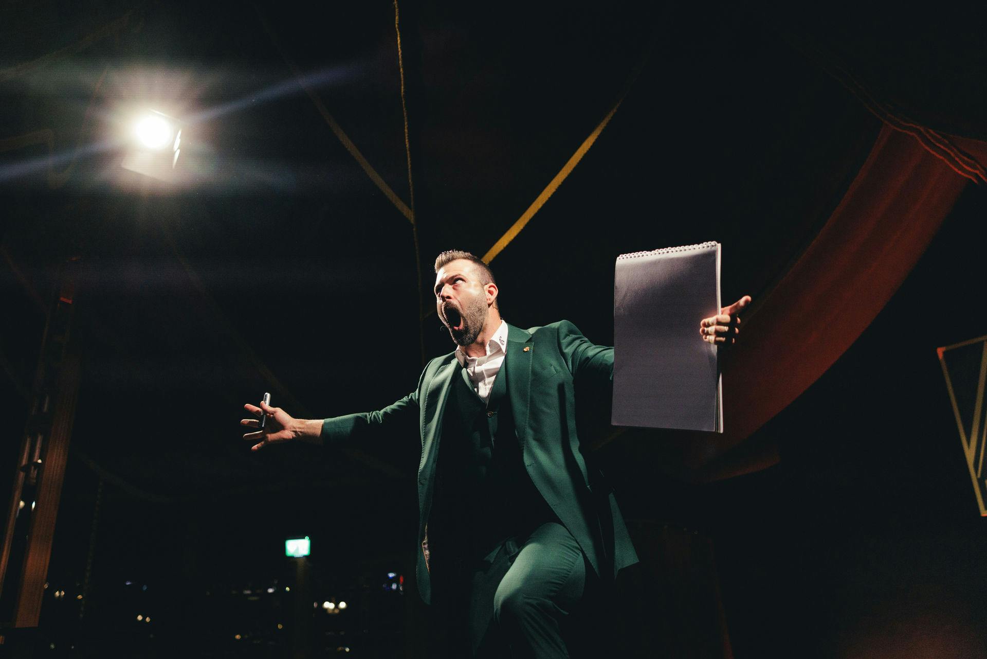 一位身穿绿色西装的人热情洋溢地说话，拿着笔记本和马克笔，在舞台灯光下。