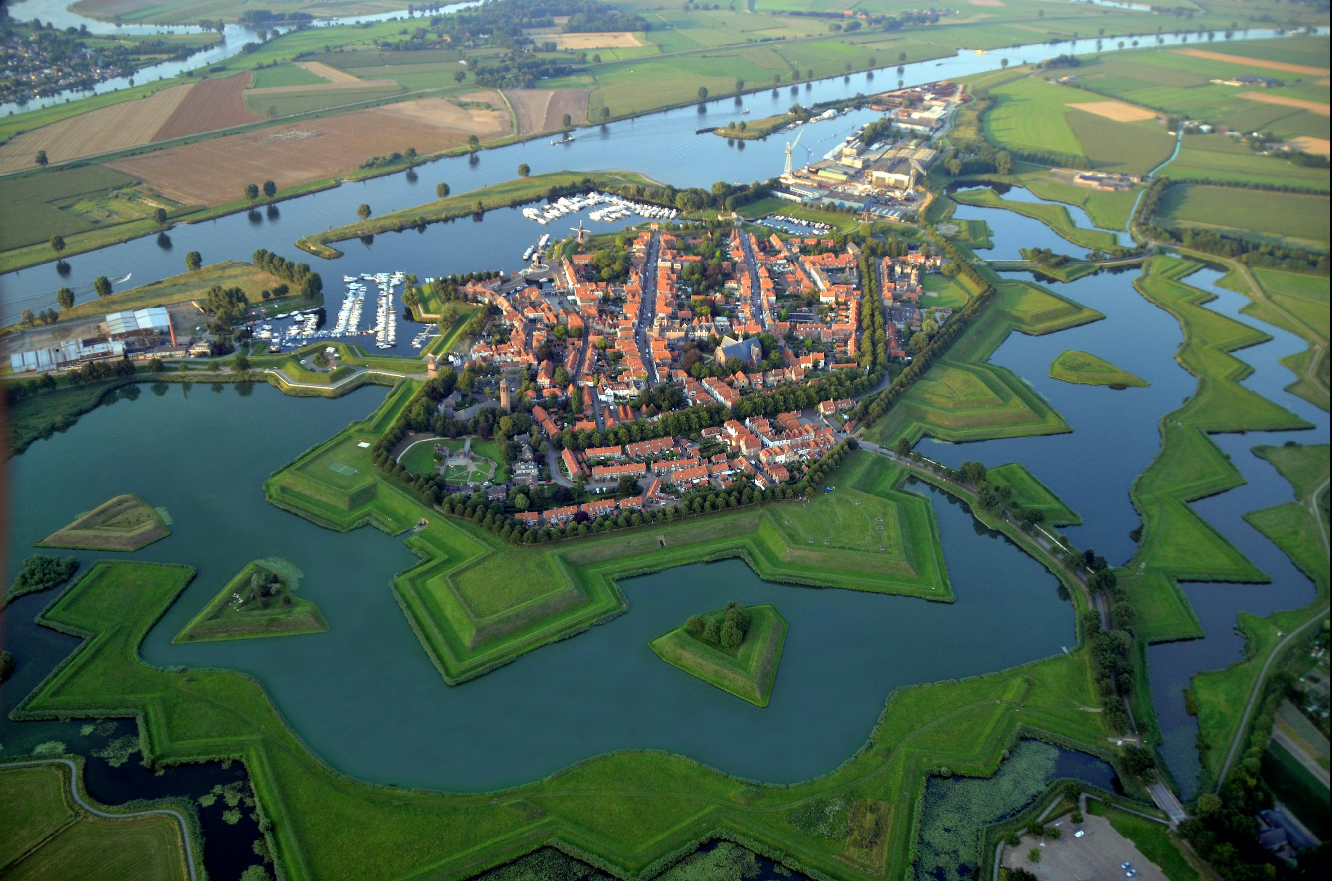 Luftfoto af en befæstet by med stjerneformede mure omgivet af vandkanaler, med bygninger med røde tage og grønne marker.
