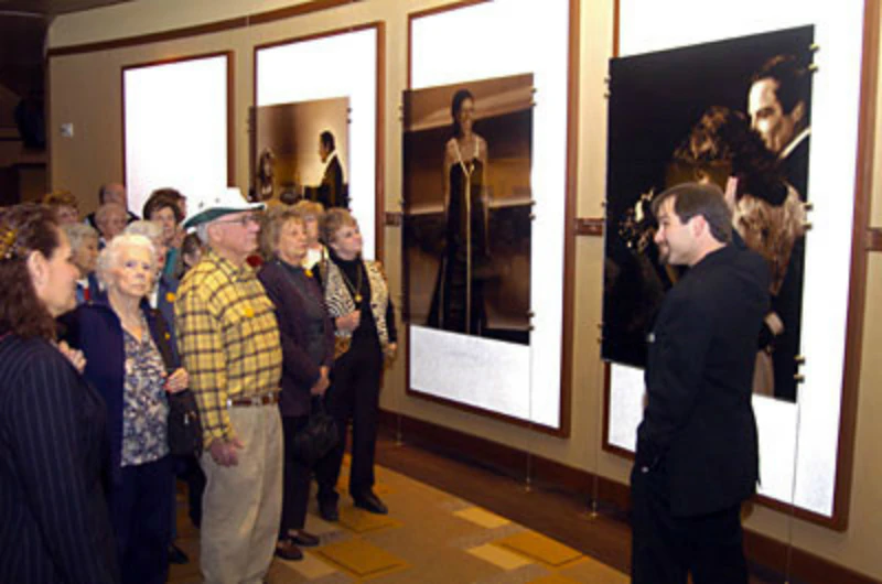 A group of elderly people stand in a museum, attentively viewing framed historical photographs of notable individuals on the wall.