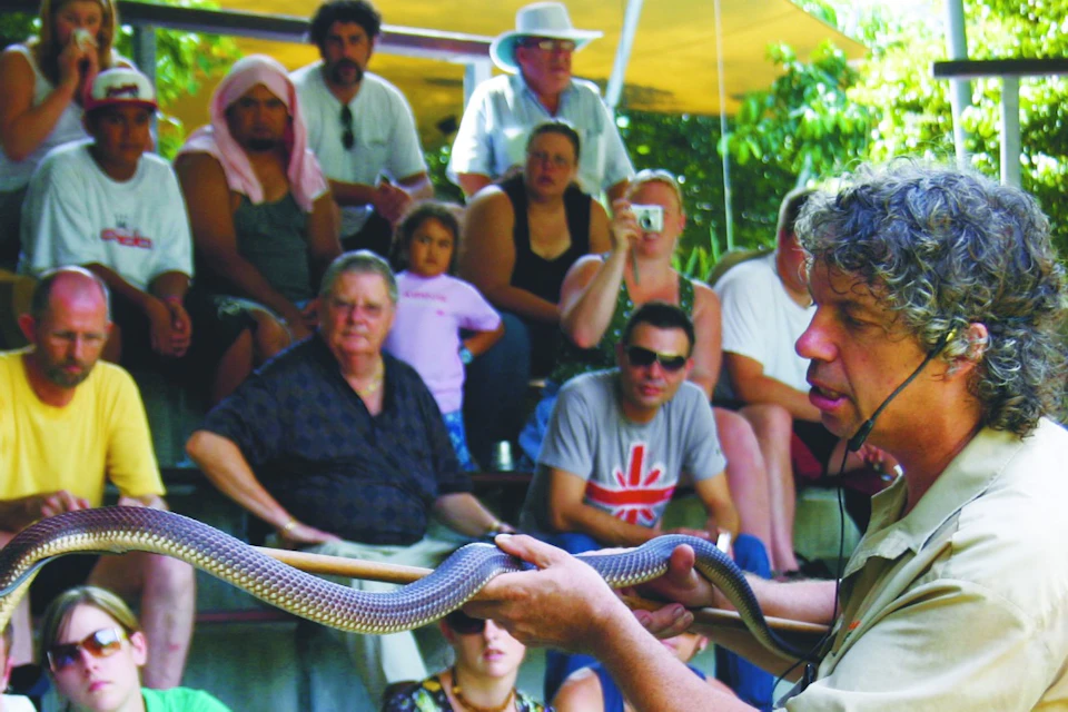 A presenter holding a snake addresses an audience seated in an outdoor amphitheater.