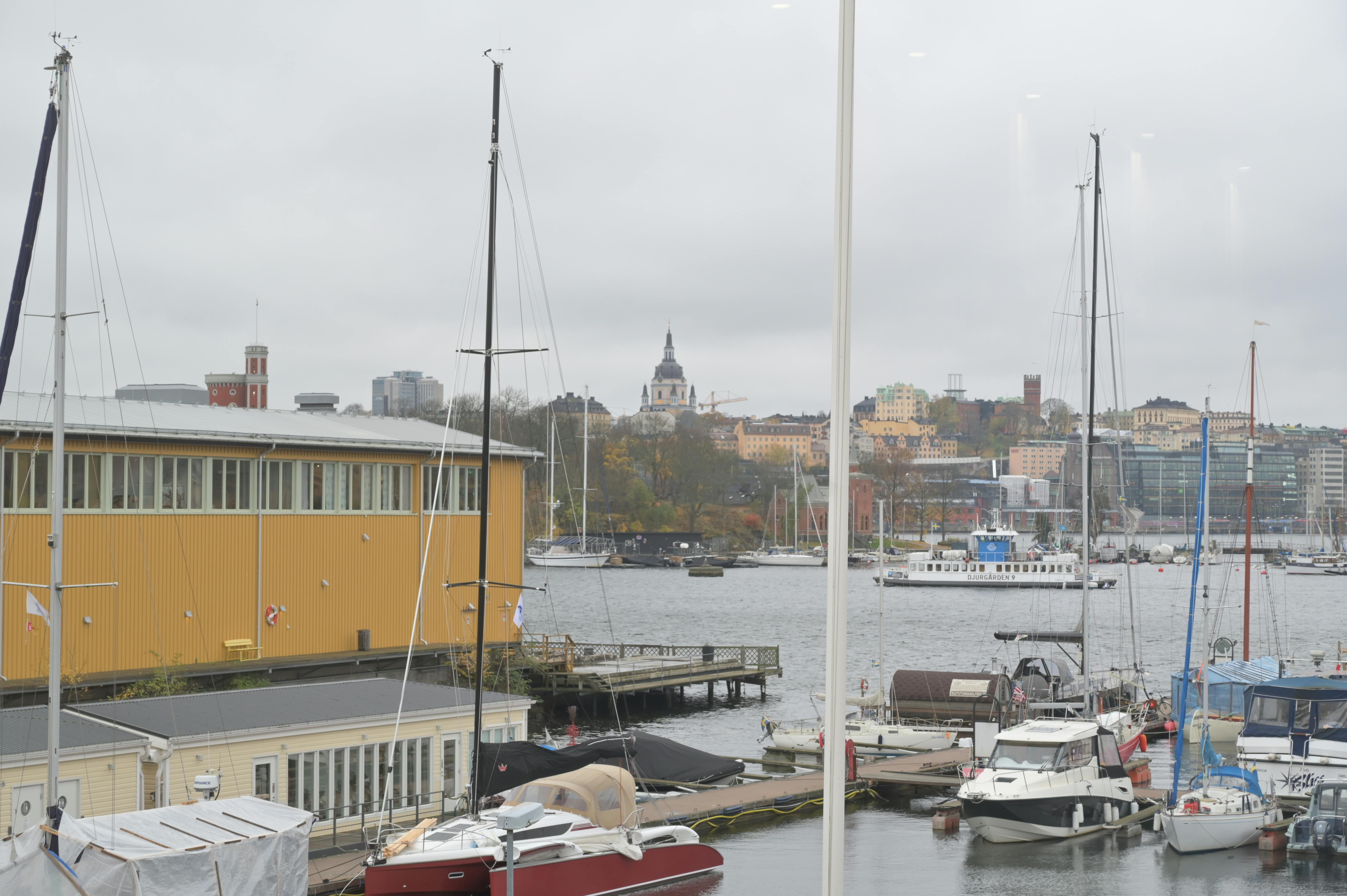 Båtar är dockade i en småbåtshamn en molnig dag, med en gul byggnad i närheten och en stadsbild i bakgrunden.