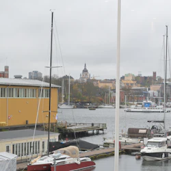 Båtar är dockade i en småbåtshamn en molnig dag, med en gul byggnad i närheten och en stadsbild i bakgrunden.