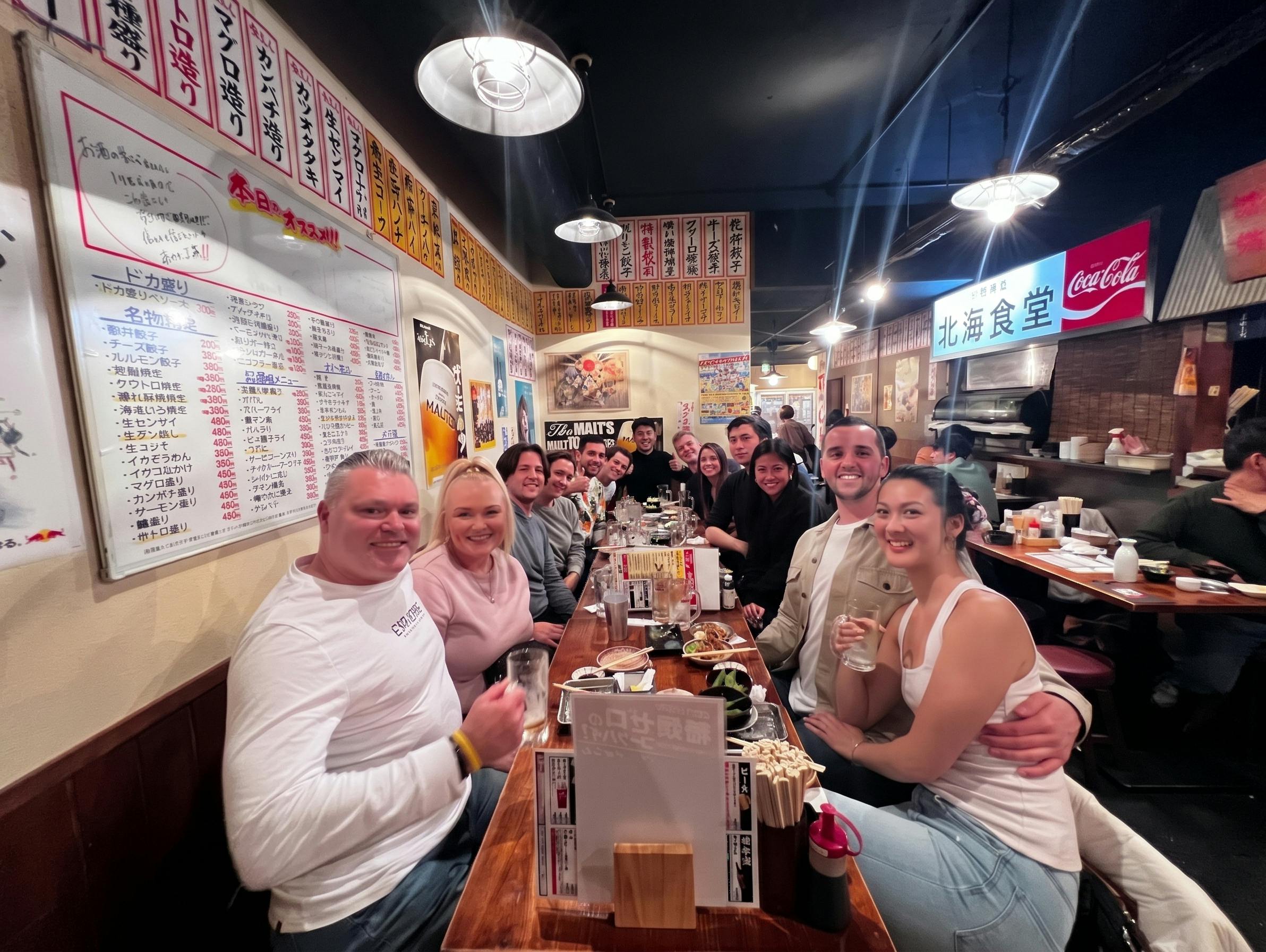 A group of people seated at a long wooden table in a bustling restaurant, smiling and enjoying food and drinks.