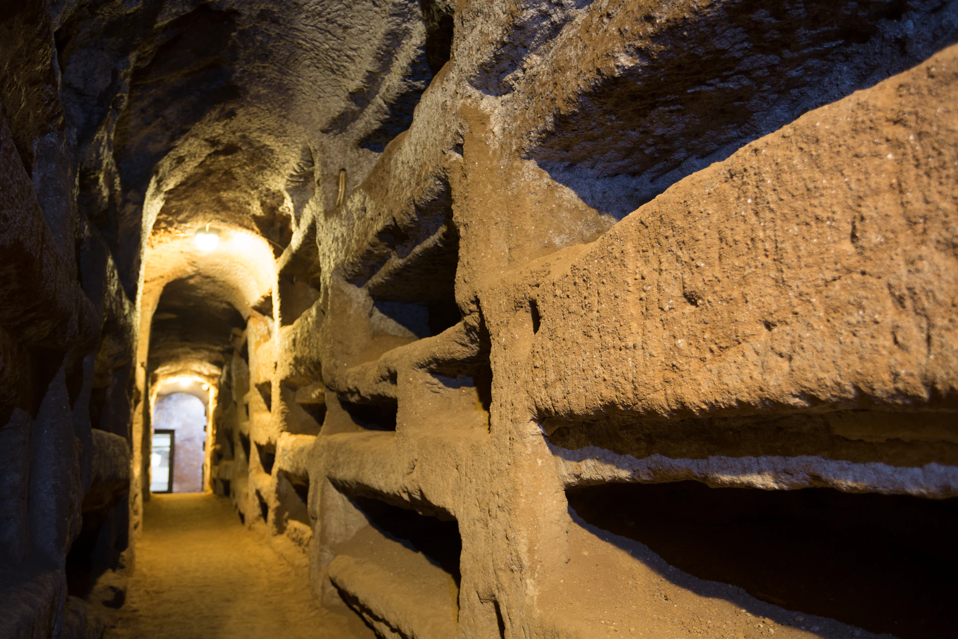 Ancient stone tunnel with arched ceilings and empty wall niches. Dim lighting illuminates the rough, carved surfaces.