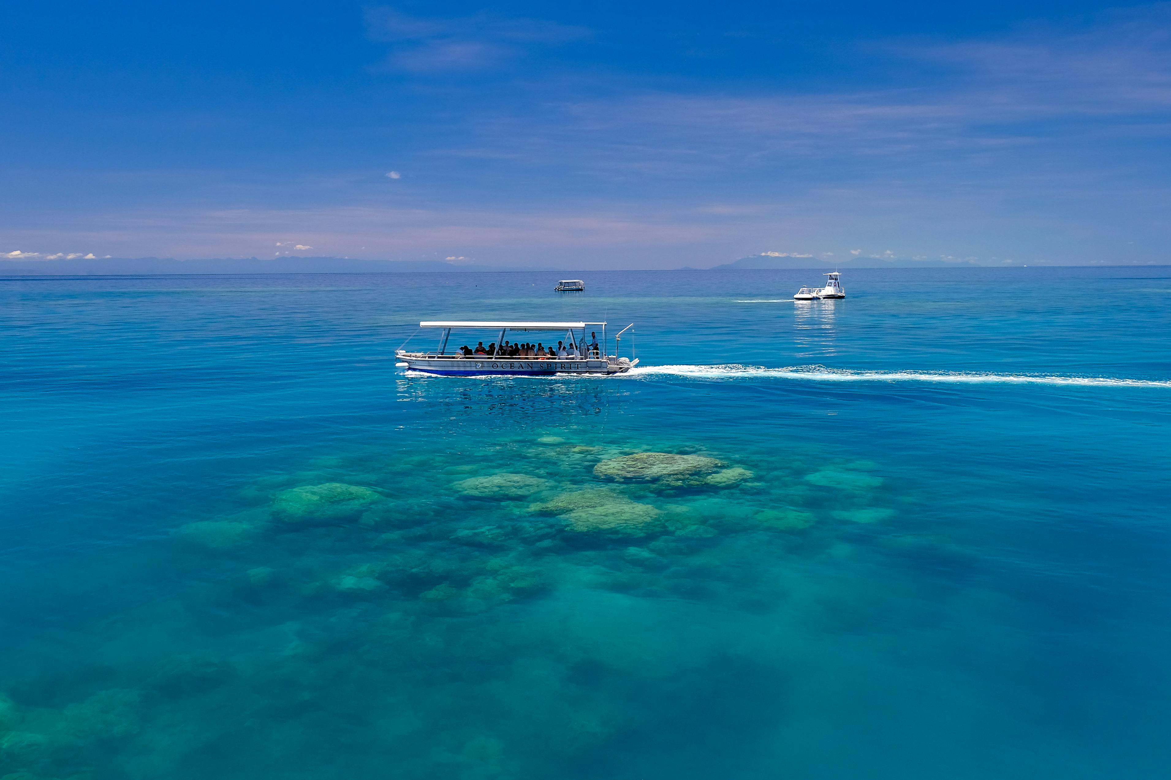Three boats sail over clear blue water with visible coral reefs below, under a sunny sky. Passengers can be seen on the nearest boat.