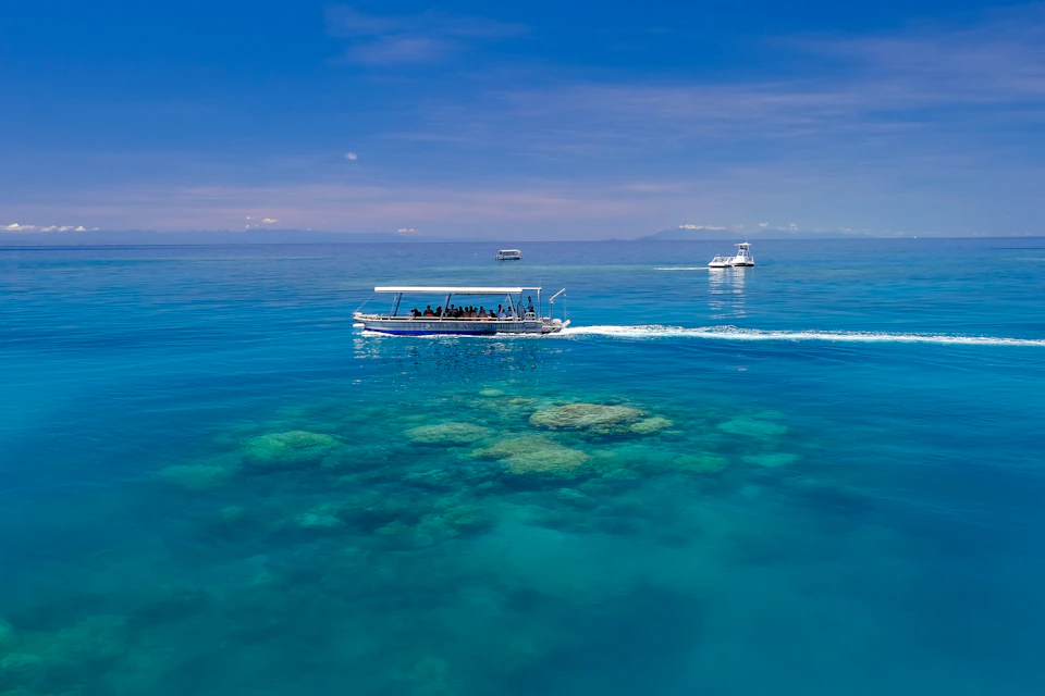Three boats sail over clear blue water with visible coral reefs below, under a sunny sky. Passengers can be seen on the nearest boat.