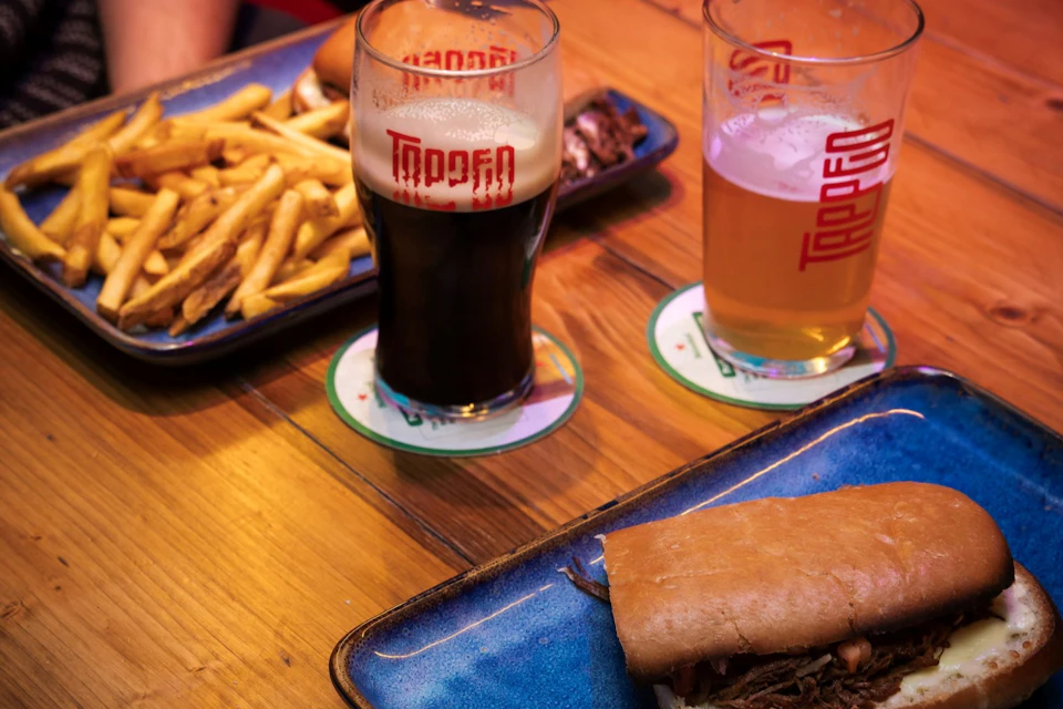 Two glasses of beer, plates with fries and a sandwich on a wooden table, served on blue dishes with branded coasters.
