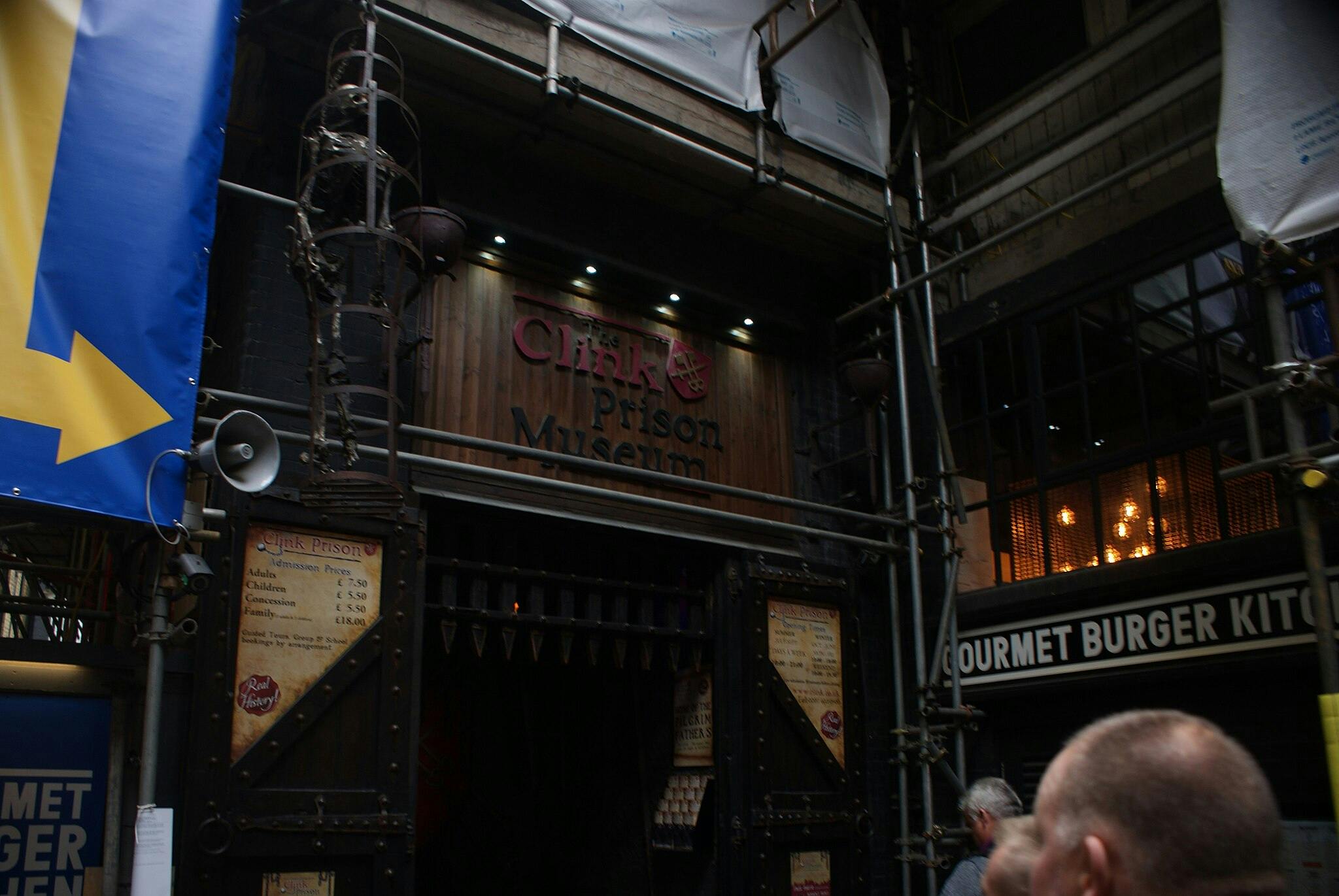 Enseigne du musée de la prison de Clink sous un échafaudage, avec une liste de prix d'entrée sur un panneau en bois. Panneau adjacent : "GOURMET BURGER KITCHEN".