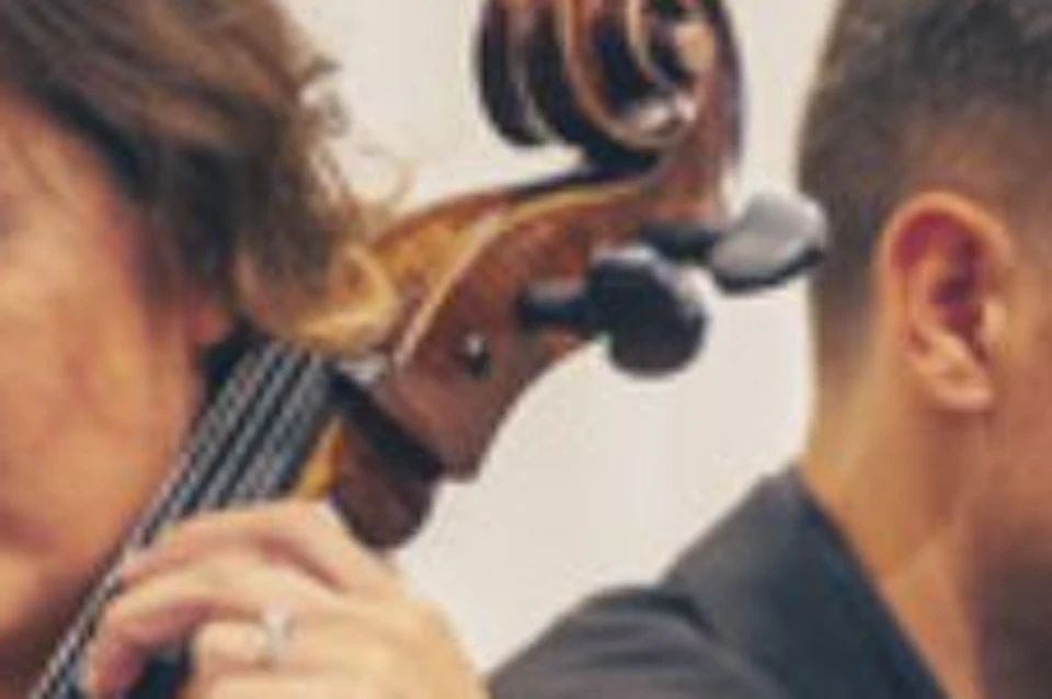 A woman in a white dress plays a cello beside a man in a black shirt playing the piano in an indoor setting.