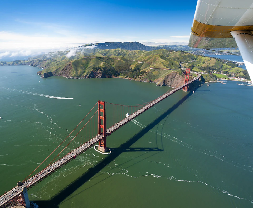 Aerial view of the Golden Gate Bridge spanning across green waters, with rolling hills and a clear blue sky in the background.