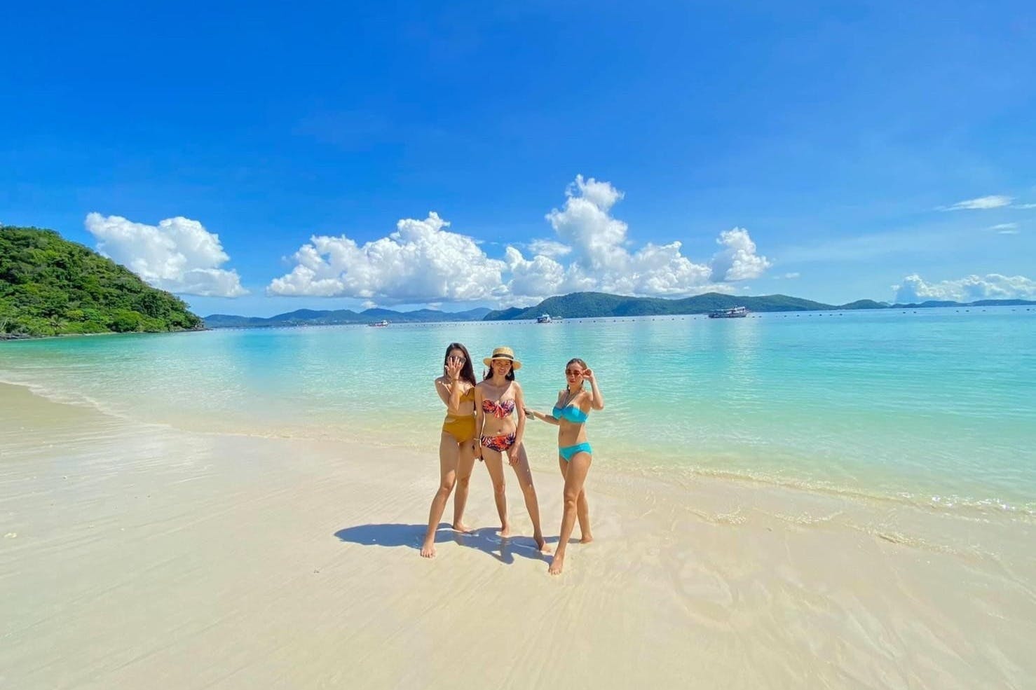 三位身着泳装的女士站在阳光明媚的海滩上，水晶般清澈，遥远的岛屿在蓬松的云层下的蓝天下。