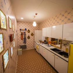 Ett smalt kök med vintage-tapeter, vita skåp, ett gult kylskåp, väggdekor, hängande redskap och inramade komiska konstverk.