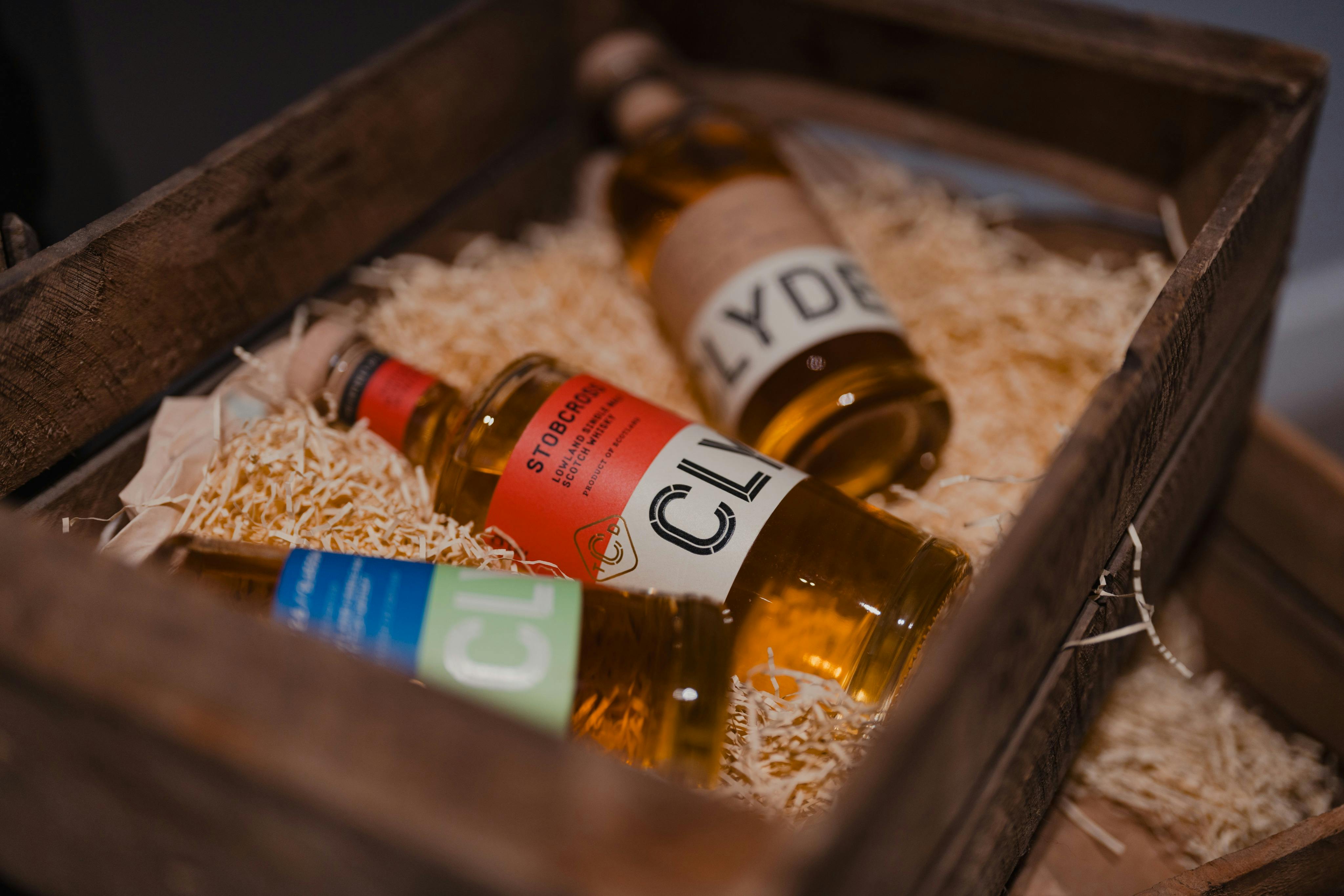 Tre flasker spiritus i en trækasse fyldt med halm, der viser farverige etiketter og korkplader.