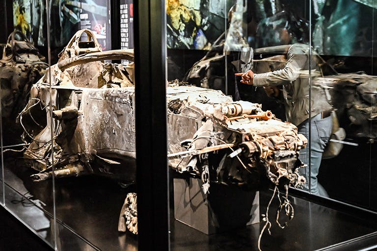 Un cotxe molt malmès i cremat es mostra en una vitrina del museu, amb reflexos de persones que el veuen.