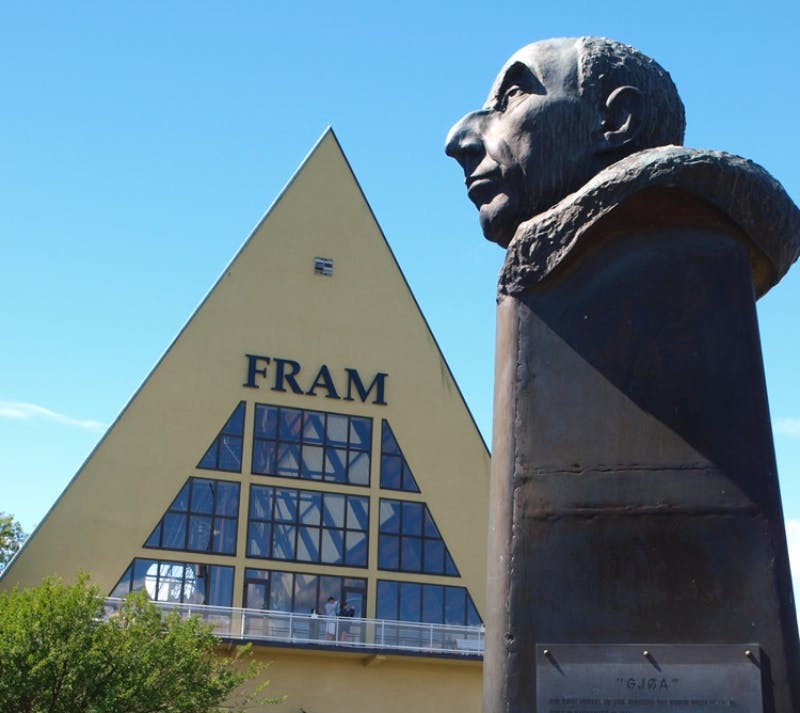 Τριγωνικό κίτρινο κτίριο με την επιγραφή «FRAM» με μεγάλα παράθυρα και την προτομή ενός άνδρα σε πρώτο πλάνο.